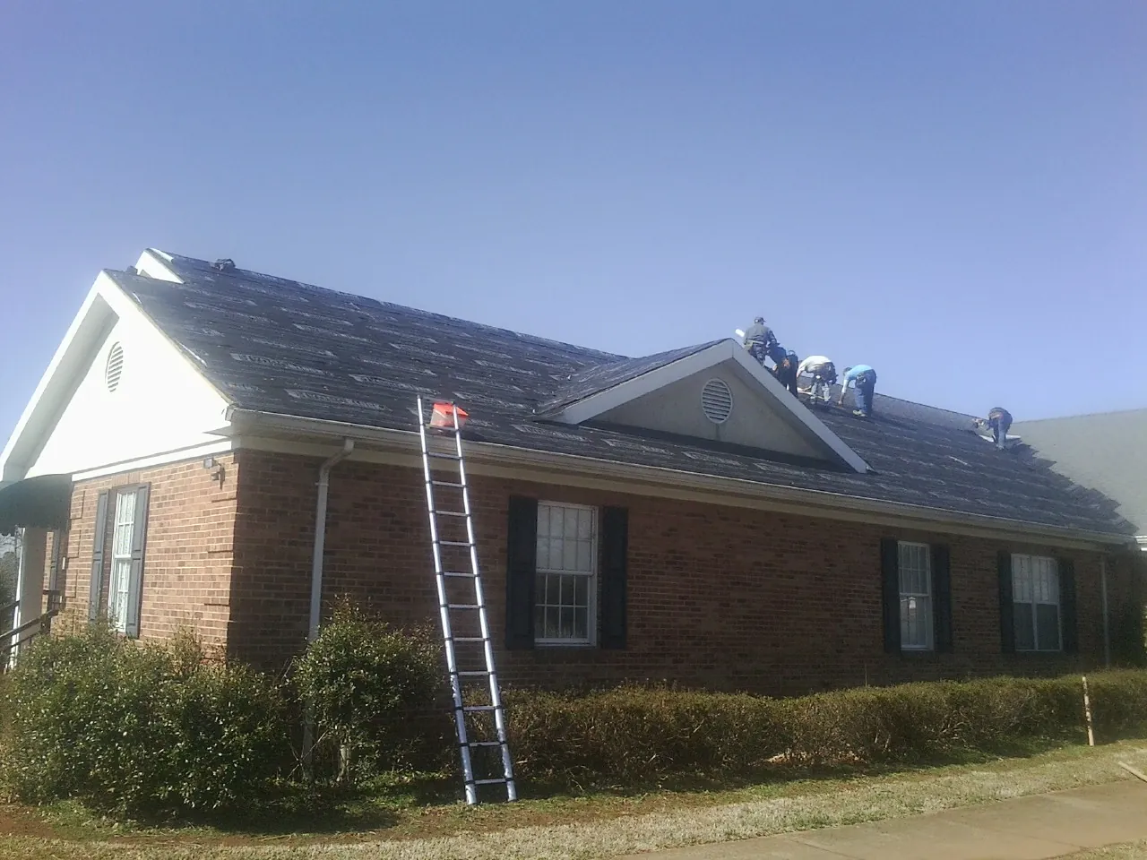 Crew tearing shingles off a brick ranch roof with a ladder set — Emergency Roof Repair work by our crew in Geneseo